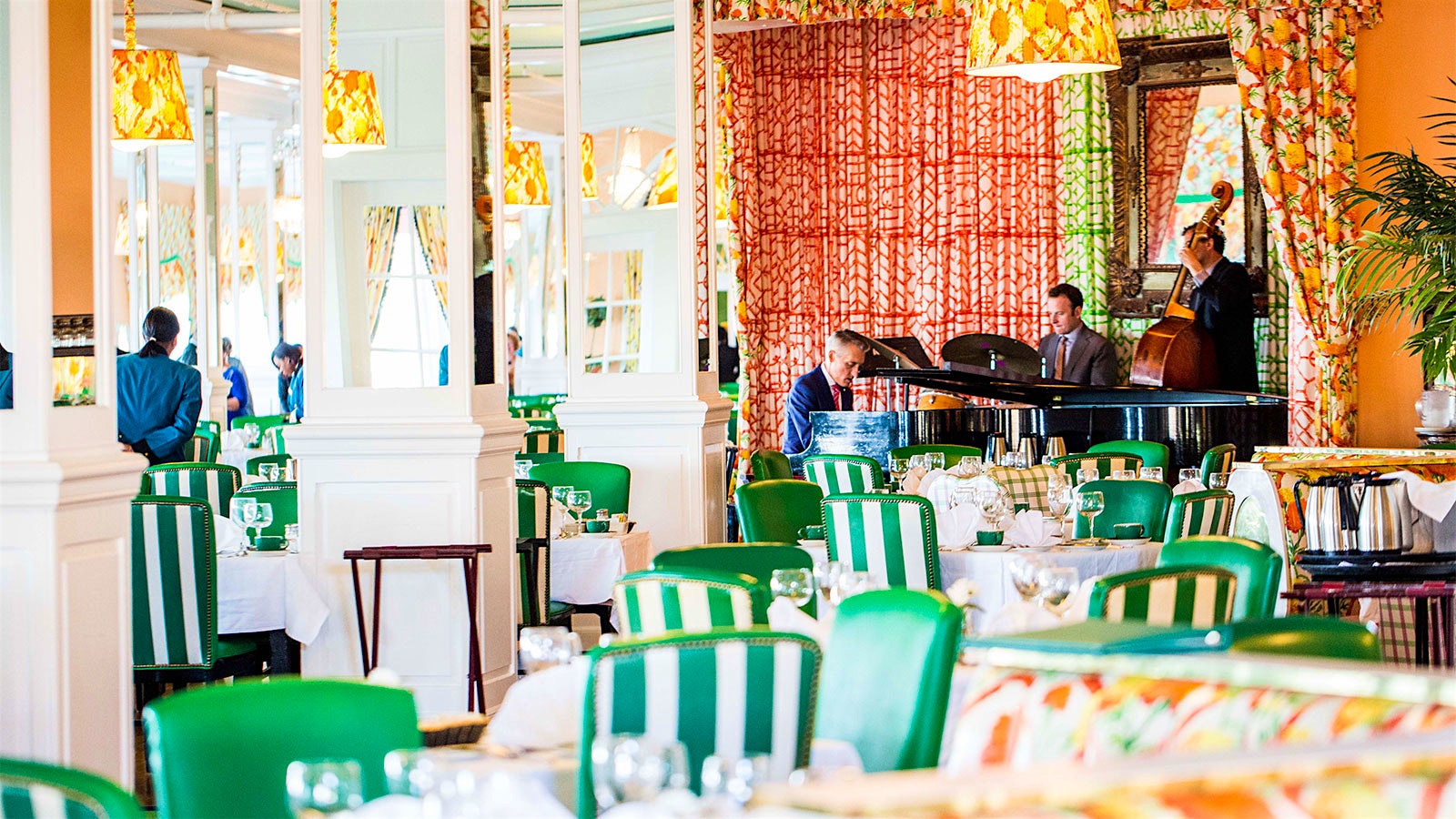 Green-and-white leather chairs and set tables in the long Main Dining Room of Grand Hotel, where a pianist, a drummer and a bassist play music